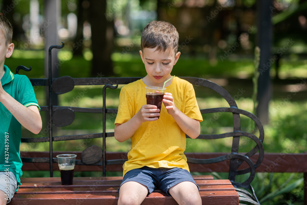 a 7-year-old boy in a yellow T-shirt drinks a cool brown drink from a glass to quench his thirst. Summer heat - obrazy, fototapety, plakaty 