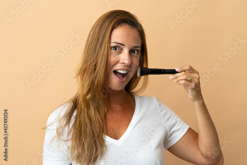 Middle age brazilian woman isolated on beige background holding makeup brush and surprised