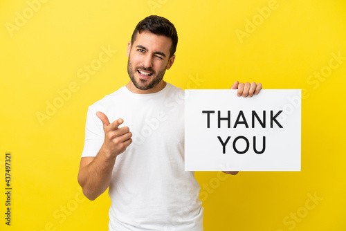 Young handsome caucasian man isolated on yellow background holding a placard with text THANK YOU and pointing to the front