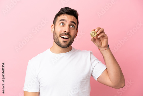 Young man holding a Bitcoin isolated on pink background thinking an idea while looking up