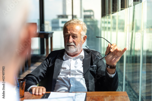 Picture of two business man discussing business and seem like an agreement is made. The young man is wearing white shirt with black tie. The older man is wearing black suit. They meet in hotel lobby.