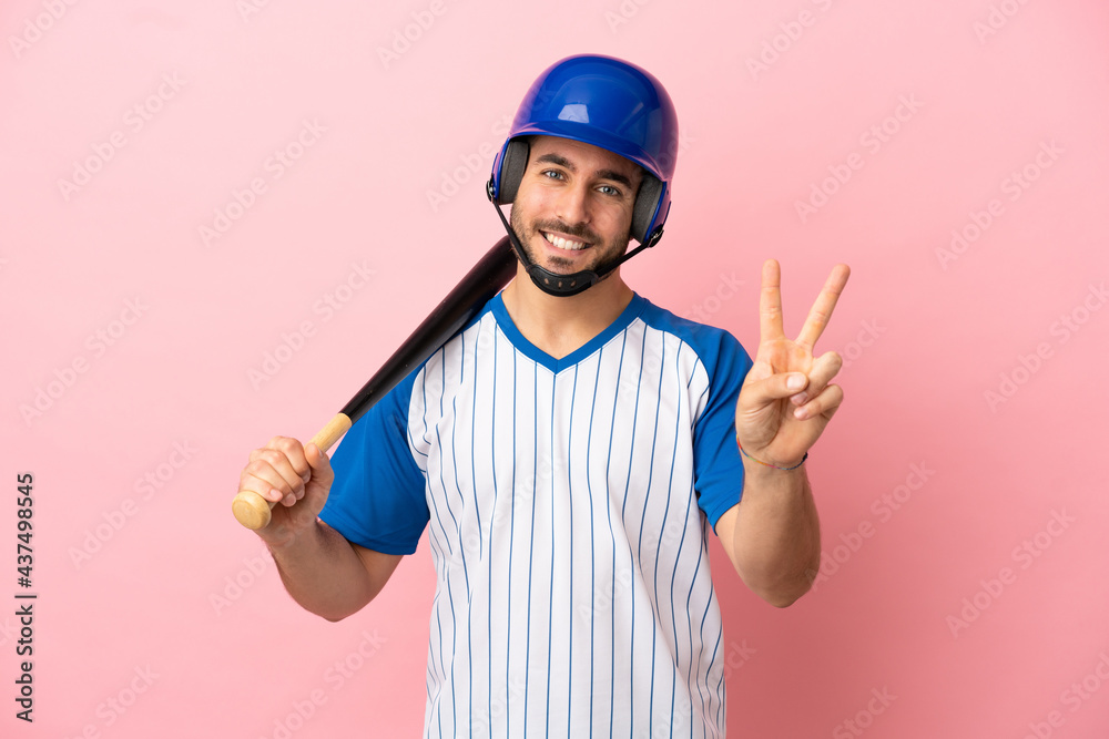 Baseball player with helmet and bat isolated on pink background smiling and showing victory sign Baseball player with helmet and bat isolated on pink background smiling and showing victory sign