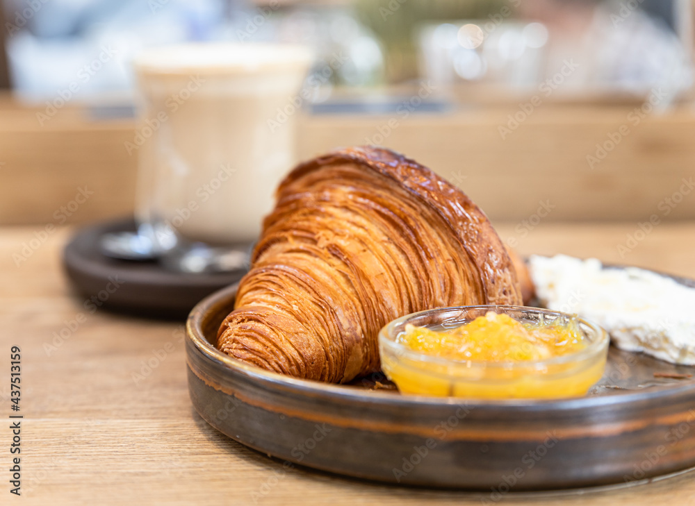 Ceramic plate with fresh crispy croissant, orange jam and butter with a glass cup of cappuccino on wooden table in cafe. Ceramic plate with fresh crispy croissant, orange jam and butter with a glass cup of cappuccino on wooden table in cafe.