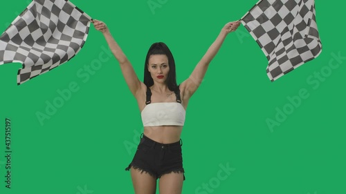 Sexy brunette in shorts waving black and white checkered racing flags to signal start of competition. Young woman posing against background of green screen close up. Slow motion ready, 4K at 59.94fps. photo