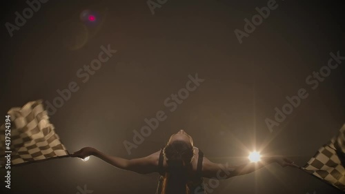 Young woman waving checkered race flags while performing in racing competition. Back view silhouette of brunette poses in dark smoky studio with backlight. Close up. Slow motion ready, 4K at 59.94fps. photo