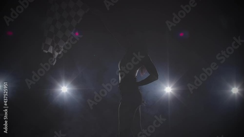 Side view of silhouette young woman waving checkered race flag to signal the start of racing event. Brunette posing in dark smoky studio with backlight. Close up. Slow motion ready, 4K at 59.94fps. photo