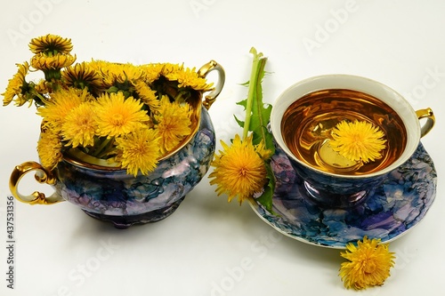 A porcelain blue vase with a bouquet of dandelions and a blue porcelain cup with dandelion tea are on a white table