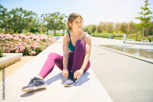 Pretty young woman laces up her sneakers sitting in a park with no people around her, getting ready for a morning workout.