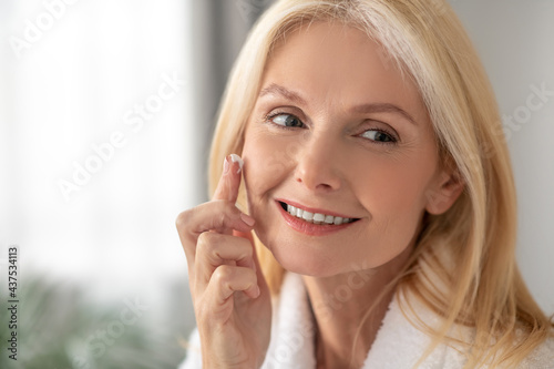 A woman in a white bath robe applying facial cream on her cheeks