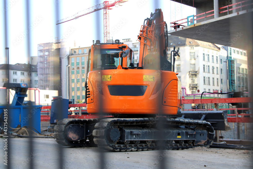 Orange crawler excavator on a construction site in the city centre behind a construction fence.