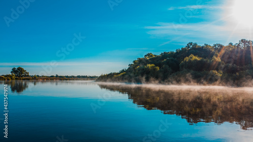 Aerial view of a beautiful summer landscape over river while dawn. Top view over river with a smooth water surface reflecting blue sky. Morning evaporation on a river while sunrise.