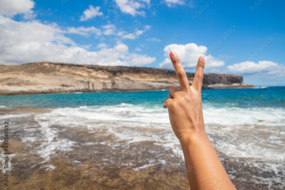 Close-up of a girl's hand making the V sign. She is at the beach enjoying the summer. Hand gesture for happiness and enjoyment concepts. Hand on focus, Blurry background.