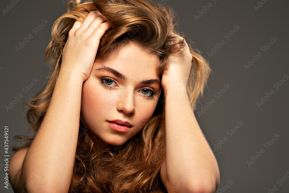 Fototapeta premium Closeup portrait of an young adult girl with long curly hair. Photo of a fashion model posing at studio. Pretty young woman with long brown hair looking at camera. Beauty portrait.