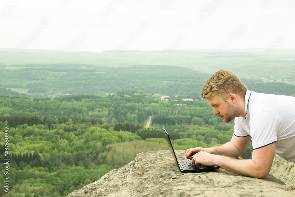 Handsome caucasian man with curly blond hair working at his notebook during trip in mountains, on background of beautiful view. Distance work, outdoor office idea