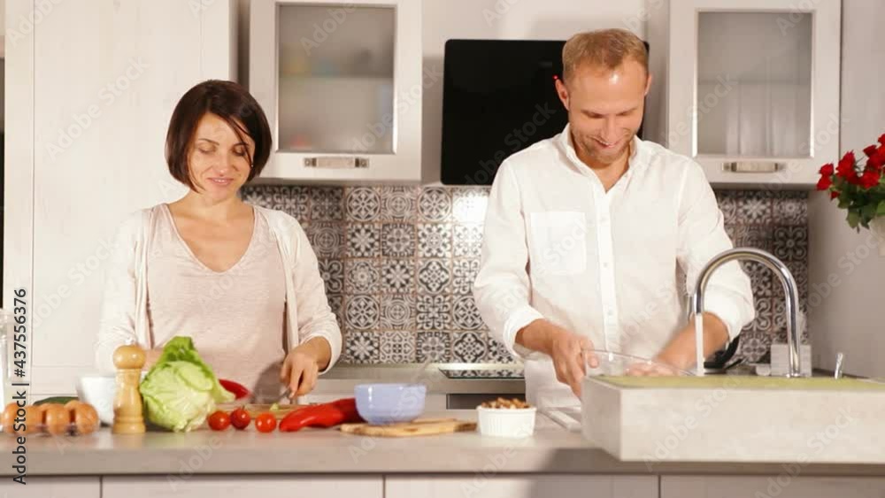 Couple in the kitchen preparing meal wearing white and talking to each other. Man washing dishes and woman rinsing vegetables to prepare salad. Healthy eating, eating at home concept. Couple in the kitchen preparing meal wearing white and talking to each other. Man washing dishes and woman rinsing vegetables to prepare salad. Healthy eating, eating at home concept.