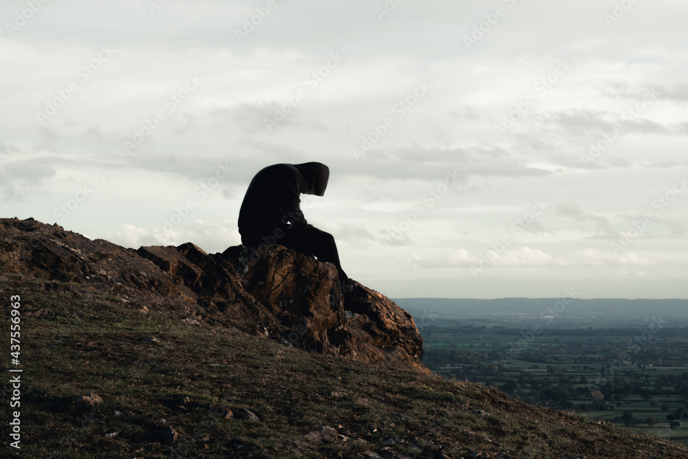 A mental health concept of a hooded man sitting alone on top of a hill in  the countryside. Looking down. Stock Photo | Adobe Stock