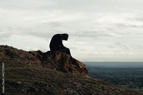 A mental health concept of a hooded man sitting alone on top of a hill in the countryside. Looking down.