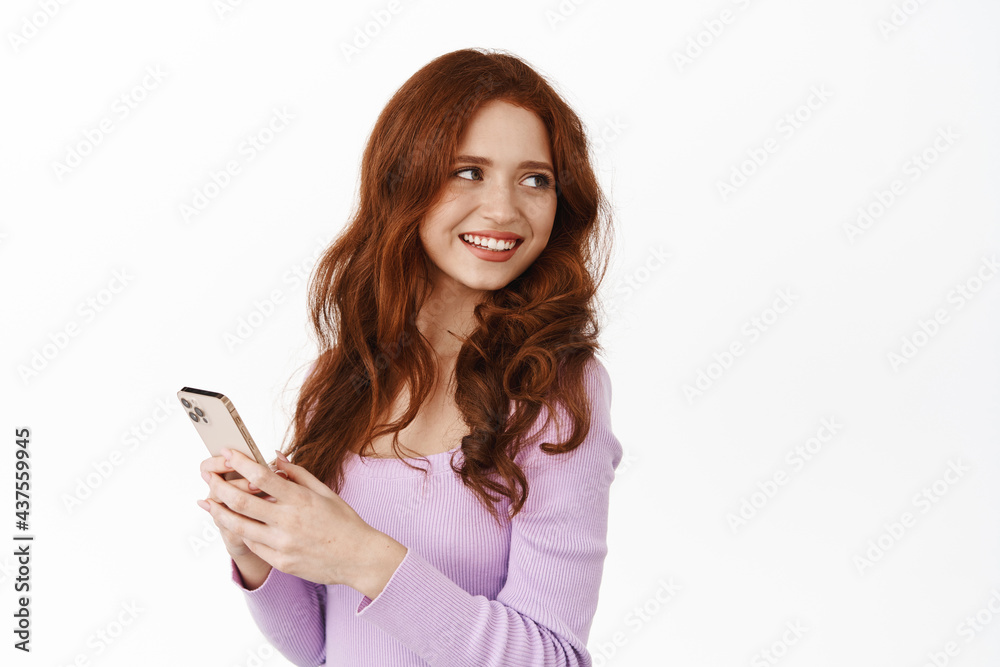 Glamour girl with ginger hair turn around to smile at promo offer, chatting on mobile phone, holding smartphone to message or use application, white background Glamour girl with ginger hair turn around to smile at promo offer, chatting on mobile phone, holding smartphone to message or use application, white background