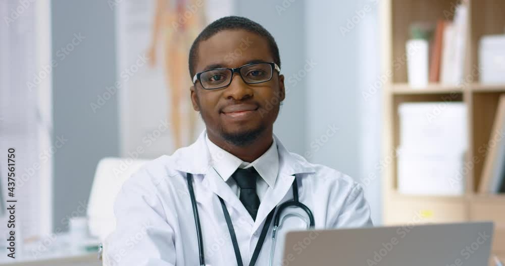 Close up portrait of young african american doctor in white coat and glasses sitting in hospital office and looking at camera smiling.