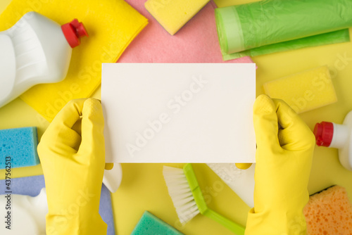 Overhead pov photo of equipment for cleaning and hands in yellow gloves with empty card isolated on the yellow background