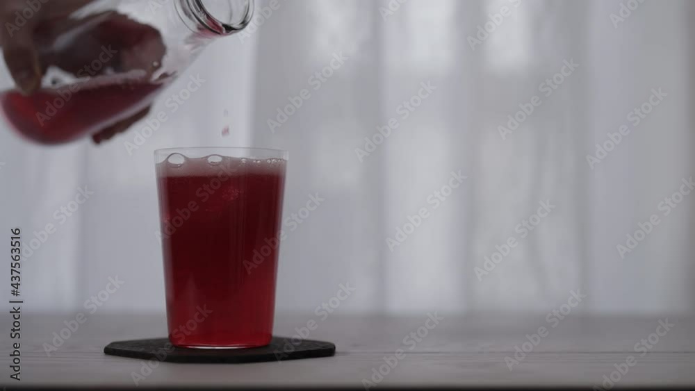 Slow motion pour red berry sparkling drink into tumbler glass with ice on white oak table Slow motion pour red berry sparkling drink into tumbler glass with ice on white oak table
