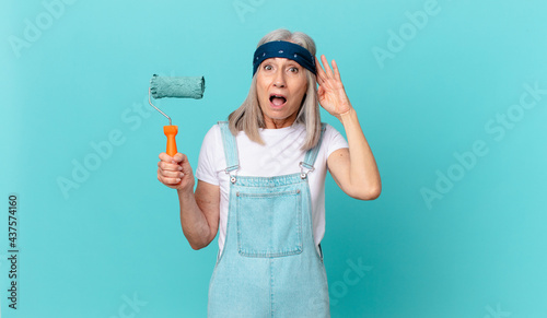 middle age white hair woman looking happy, astonished and surprised with a roller painting a wall