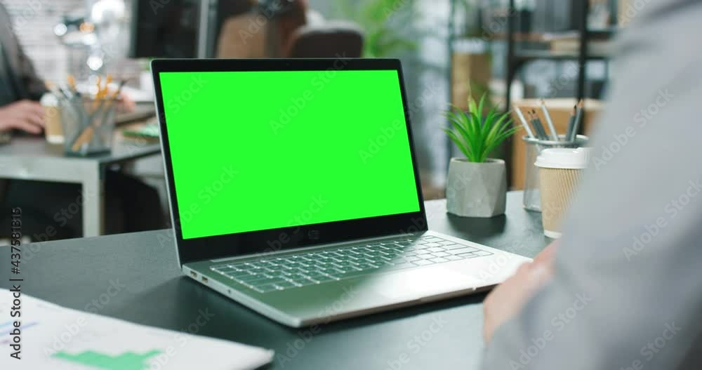 Over the shoulder: creative young man sitting at his desk using laptop computer with mock-up green screen. Bright office where diverse team of young professionals working on computers