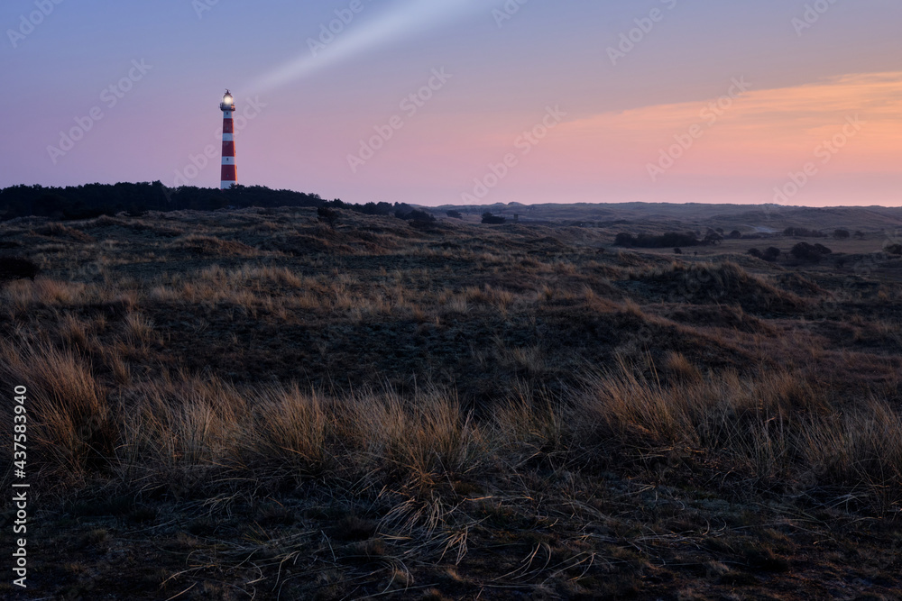 Historic lighthouse of Hollum with light beam, Ameland with a red and orange sky during sunset, sunrise. Dark dunes with stern grass in the foreground.