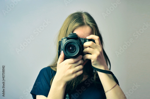 Girl with a SLR camera in her hands. Front view. The camera lens is directed towards the viewer. The camera completely covers the face. A girl of European appearance with long hair and a blue T-shirt.