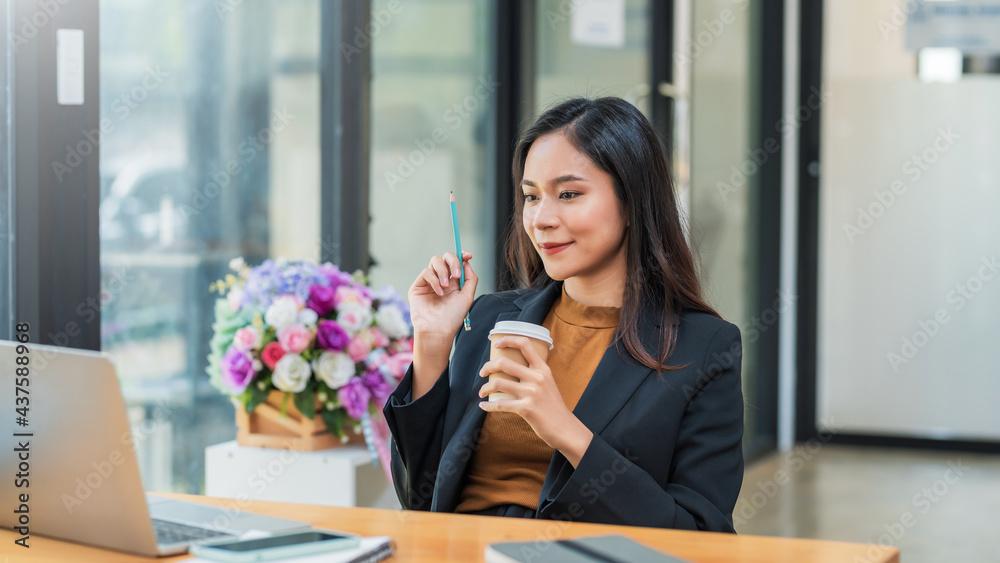 Charming Asian businesswoman holding a pencil and drinking coffee on a leisurely day on the laptop placed at the office table. Charming Asian businesswoman holding a pencil and drinking coffee on a leisurely day on the laptop placed at the office table.