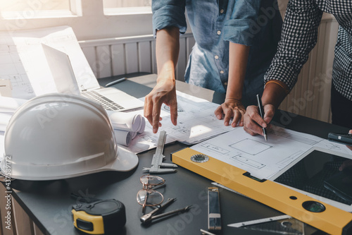 Close-up of architect and engineers holding a pen working on blueprints with safety equipment placed at the office.