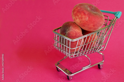 on the right, three pink whole fig peaches lie in a mini trolley from a supermarket on a pink background. side view. artificially derived fruit rich in vitamins photo