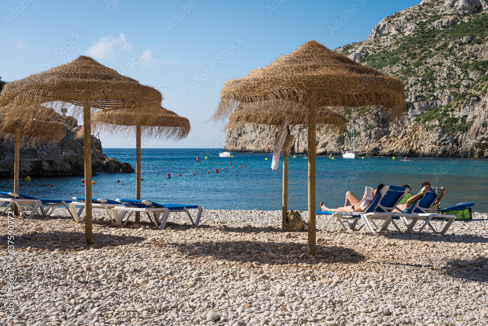 Umbrellas and hammocks on a small pebble beach on the coast of Javea on a sunny summer day, Spain