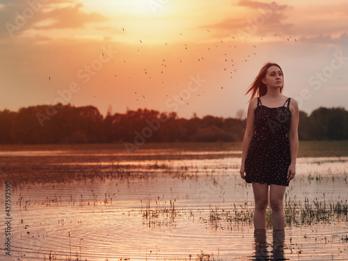 A mysterious portrait of a young woman in a short dress at sunset. Standing in the water, a flock of birds in the sky
