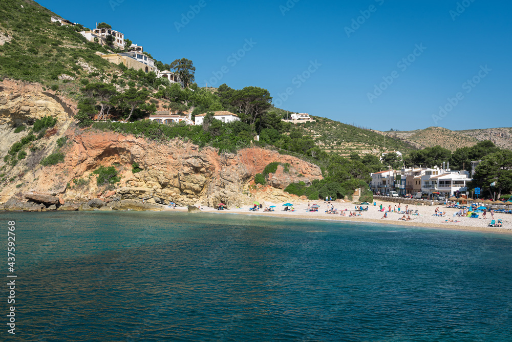 Small pebble beach at the foot of the mountain on the coast of Javea with people sunbathing on a sunny summer day, Spain