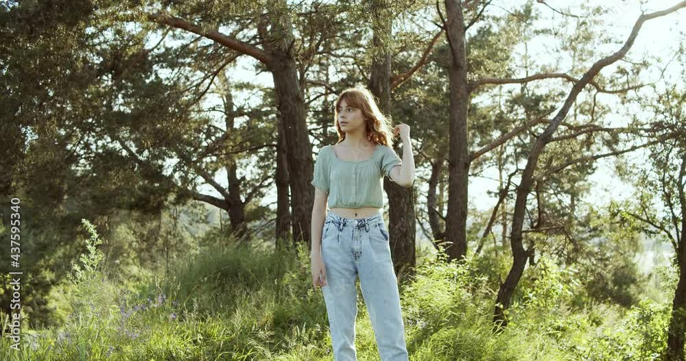 A cheerful teenage girl with curly hair stands in a clearing between green trees in summer forest and has a pleasure A cheerful teenage girl with curly hair stands in a clearing between green trees in summer forest and has a pleasure