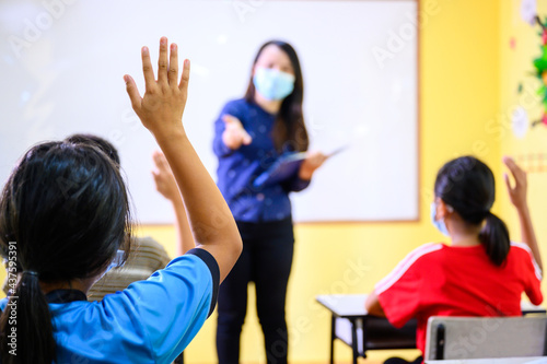 Asian female teacher Wearing a mask to prevent coronavirus (COVID 19) teaching elementary school students in a rural school and students raise their hands to answer the teacher's questions.
