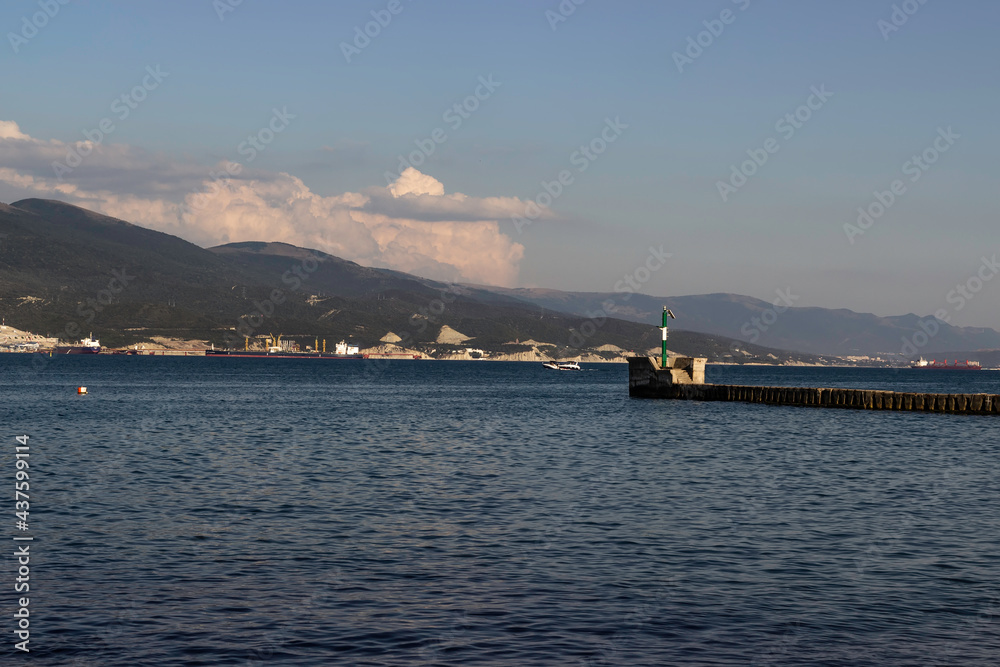 A windsurfer sails near the lighthouse against the backdrop of mountains in a sea bay on a clear day.