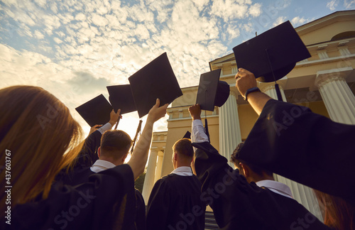 Graduation ceremony event celebration. Diverse student wearing gown mantles standing in group holding raised hat rear view from bottom. Shot over sunset and university high school building photo