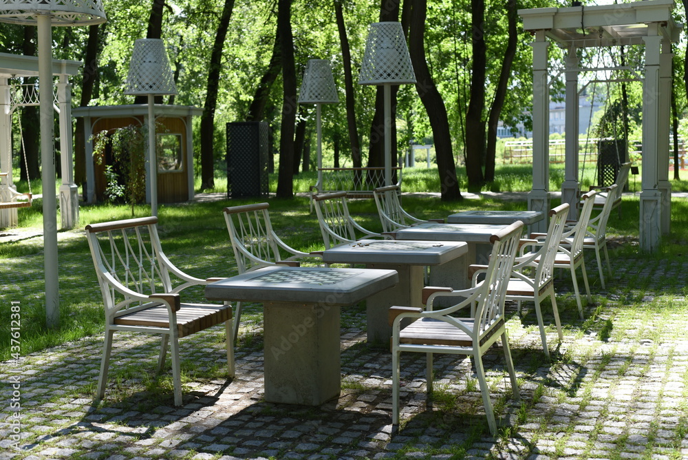 In the summer park, on a leisure area surrounded by greenery, there are empty chess tables and chairs, swing benches, lampshades in white color in an exquisite design.