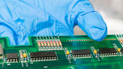 Electronic components on green printed circuit board on human hand in blue glove. Closeup of black IC chips, capacitors or resistors with standard color code on PCB detail of inside computer keyboard. photo