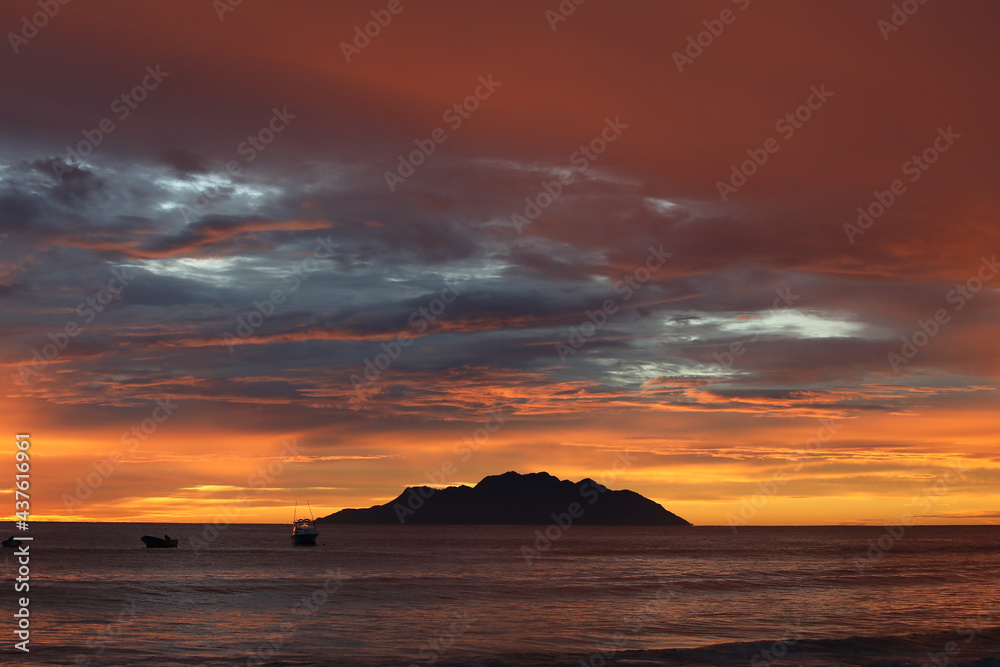 An orange glow in the sky with glimpses of a silvery cloud in the sky and the silhouette of an island on the horizon.Fiery sunset landscape at sea over a lagoon with fishing boats.Warm night image