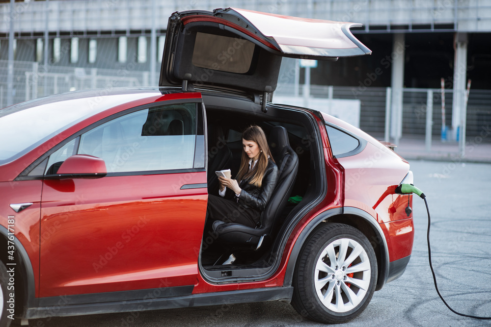 Charming woman sitting inside charging electric car with modern smartphone and cup of coffee in hands. Caucasian female talking by video call while waiting her auto to be charged.