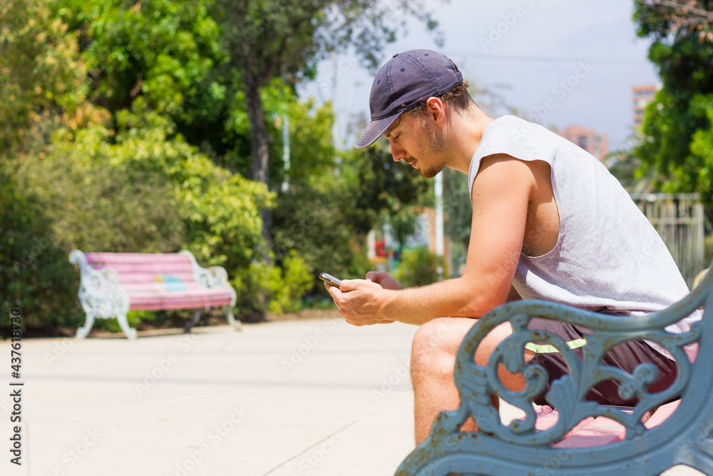 Fototapeta premium Young man holding cellphone sitting on park bench on sunny day. Boy on sleeveless shirt and shorts using smart phone outdoors in summer time