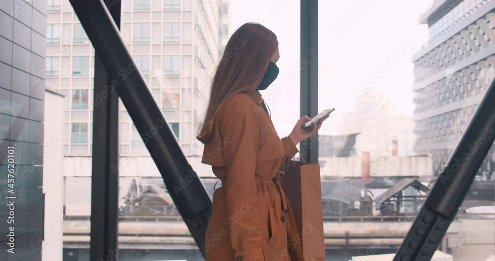 Working on the go. Side view young beautiful business woman in face mask using mobile app at mall with shopping bags. Working on the go. Side view young beautiful business woman in face mask using mobile app at mall with shopping bags.