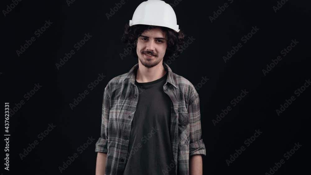 The charismatic hipster with a white engineer's helmet on his head flirts with eyebrows and looks forward proudly. Isolated on black background. The concept of life. People's emotions. 4k portrait The charismatic hipster with a white engineer's helmet on his head flirts with eyebrows and looks forward proudly. Isolated on black background. The concept of life. People's emotions. 4k portrait