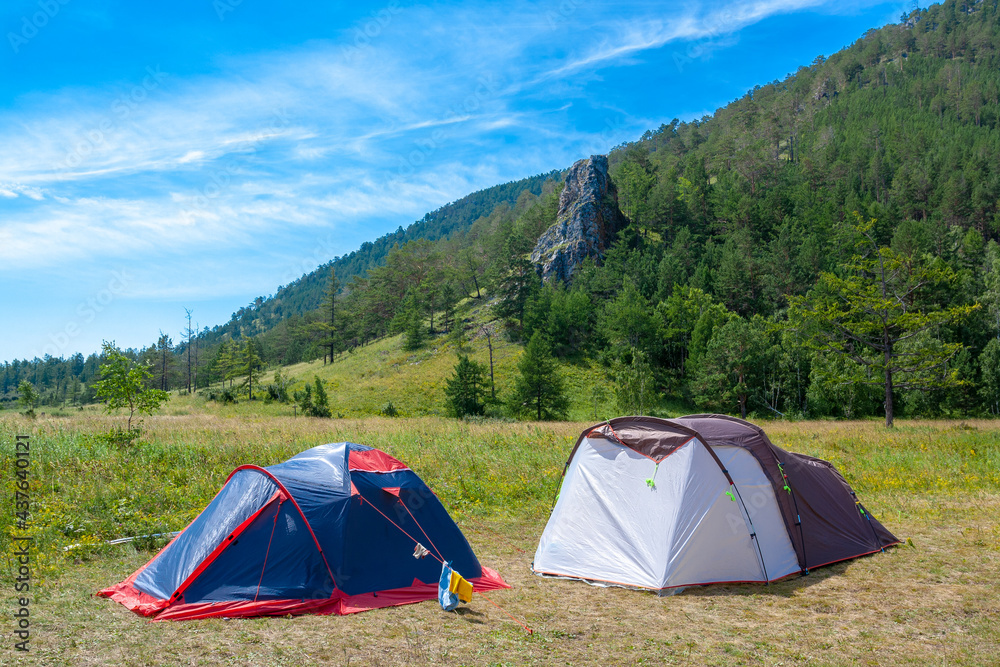 Two open tents stand on grass in the mountains. Big rock and blue sky on background. Hiking camp.