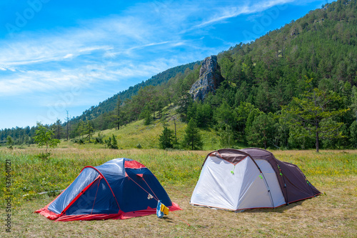 Two open tents stand on grass in the mountains. Big rock and blue sky on background. Hiking camp. © Vladimir Kazakov