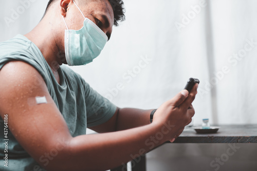 Youngman wearing a protective mask Show plaster on the shoulder Use your smartphone to find information about the impact of post-vaccination against covid-19, health care guidelines, health protection