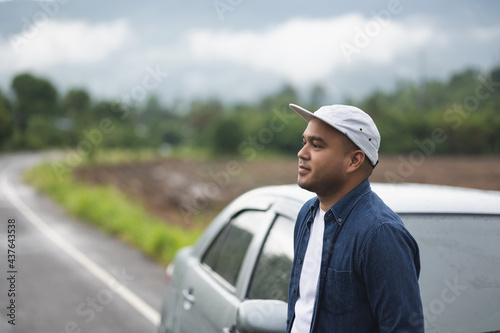A young Asian man drives a beautiful nature drive. He was standing in front of the car on the roadside.